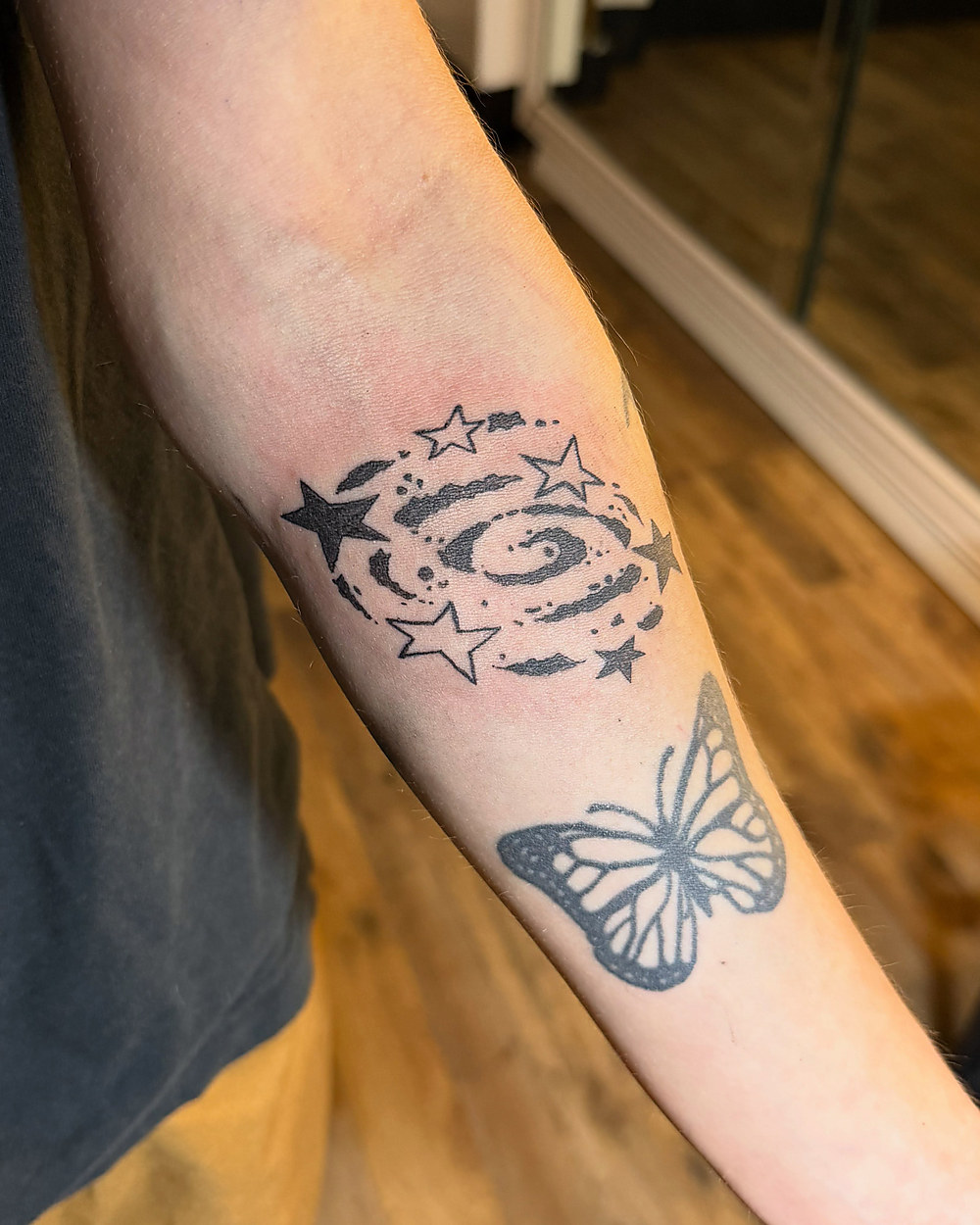 Stars, a spiral and a butterfly in fine line down a forearm, by Miah
