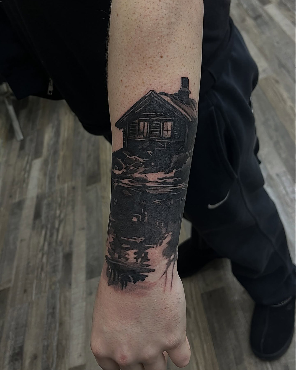 Lakeside cabin scene on a forearm, by Owen Lewis
